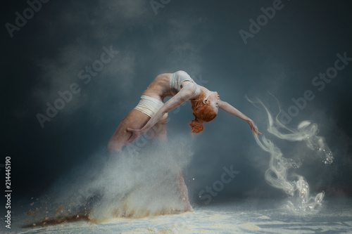 Dancing in flour concept. Redhead beauty female girl adult woman dancer in dust / fog. Girl wearing white top and shorts making dance element in flour cloud on isolated grey black background