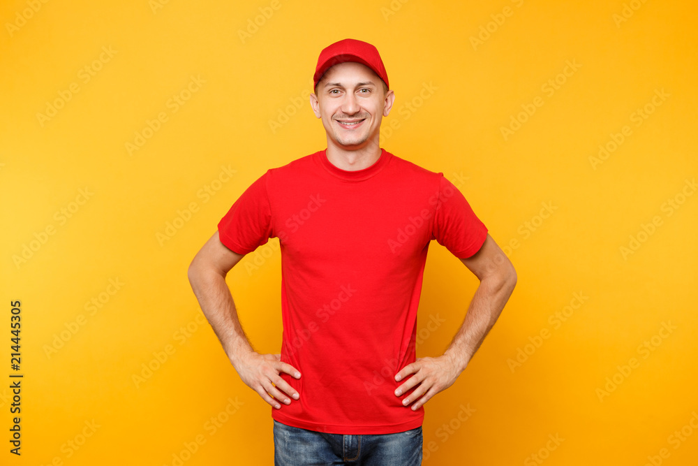 Delivery man in red uniform isolated on yellow orange background. Professional smiling male employee in cap, t-shirt working as courier or dealer standing with arms akimbo. Service concept. Copy space Delivery man in red uniform isolated on yellow orange background. Professional smiling male employee in cap, t-shirt working as courier or dealer standing with arms akimbo. Service concept. Copy space