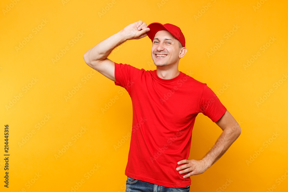 Delivery man in red uniform isolated on yellow orange background. Professional smiling male employee in t-shirt working as courier or dealer put hand on cap head gesture. Service concept. Copy space Delivery man in red uniform isolated on yellow orange background. Professional smiling male employee in t-shirt working as courier or dealer put hand on cap head gesture. Service concept. Copy space