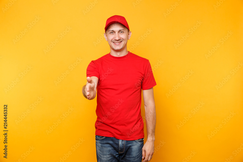 Delivery man in red uniform isolated on yellow orange background. Smiling male employee in cap, t-shirt working as courier dealer standing with outstretched hand for greeting gesture. Service concept.