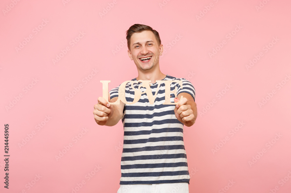 Portrait of emotional young man in striped t-shirt hold wooden word Love isolated on trending pastel pink background. People sincere emotions lifestyle holiday concept. Advertising area for copy space Portrait of emotional young man in striped t-shirt hold wooden word Love isolated on trending pastel pink background. People sincere emotions lifestyle holiday concept. Advertising area for copy space
