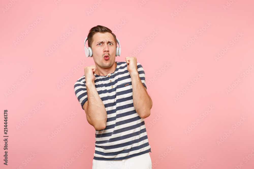Portrait of smiling young man wearing striped t-shirt listening to music with white wireless headphones, enjoy isolated on trending pastel pink background. People sincere emotions lifestyle concept. Portrait of smiling young man wearing striped t-shirt listening to music with white wireless headphones, enjoy isolated on trending pastel pink background. People sincere emotions lifestyle concept.