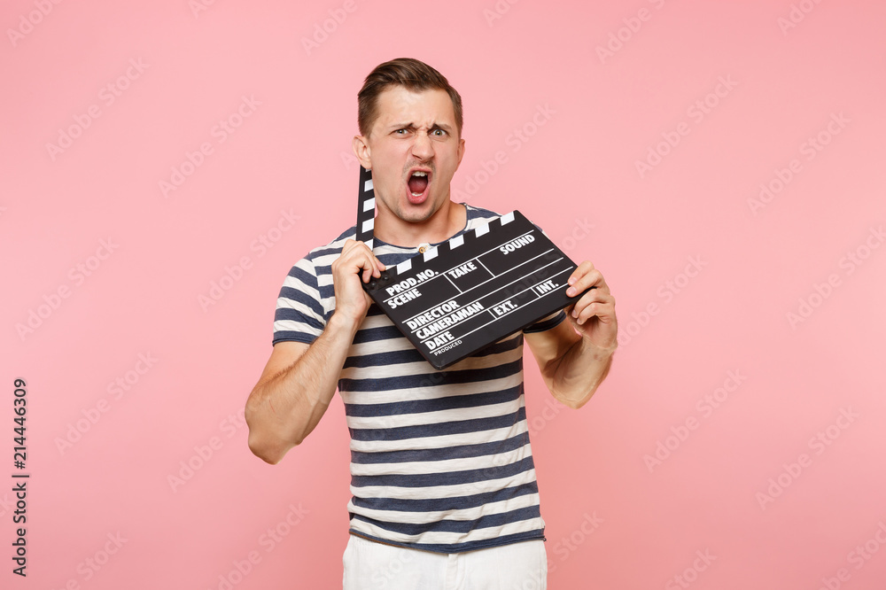Portrait of young man wearing striped t-shirt holding classic black film making clapperboard isolated on trending pastel pink background. People sincere emotions lifestyle concept. Advertising area. Portrait of young man wearing striped t-shirt holding classic black film making clapperboard isolated on trending pastel pink background. People sincere emotions lifestyle concept. Advertising area.