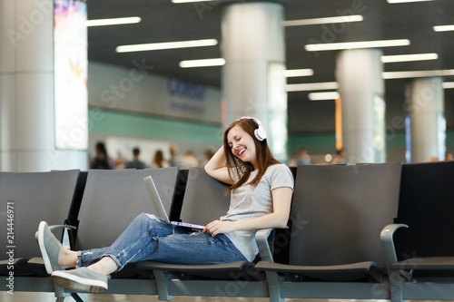 Young joyful traveler tourist woman with headphones listening music working on laptop, wait in lobby hall at international airport. Passenger traveling abroad on weekend getaway. Air flight concept.