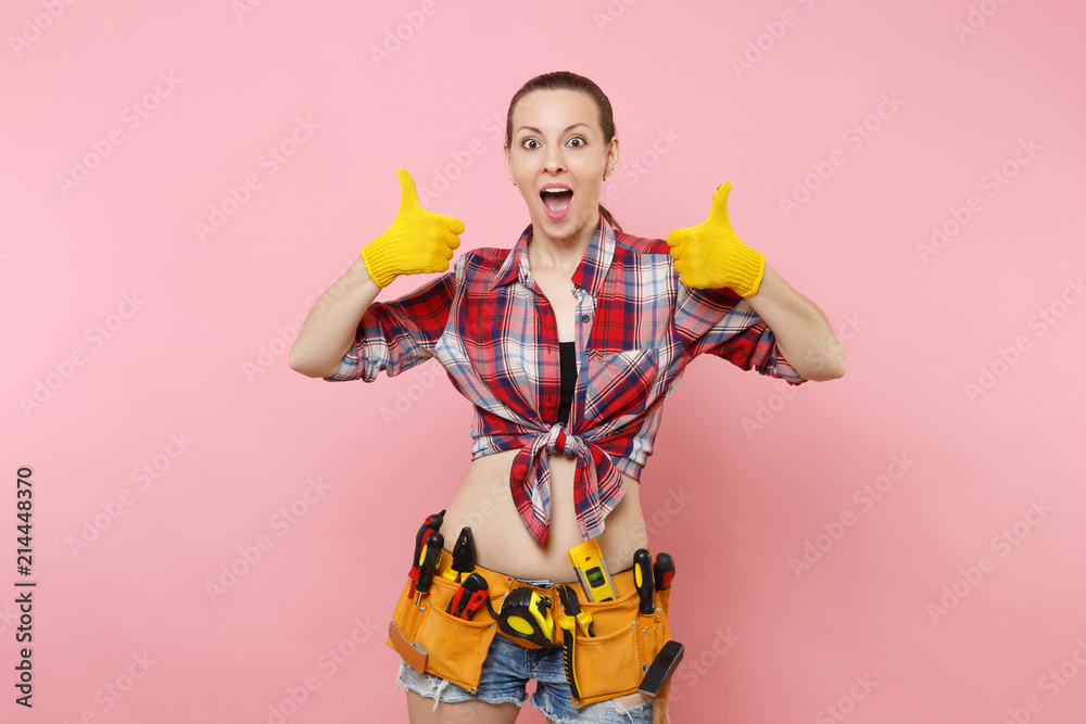 Happy handyman woman in shirt, denim shorts, yellow gloves, kit tools belt full of variety useful instruments showing thumbs up isolated on pink background. Female in male work. Renovation concept. Happy handyman woman in shirt, denim shorts, yellow gloves, kit tools belt full of variety useful instruments showing thumbs up isolated on pink background. Female in male work. Renovation concept.