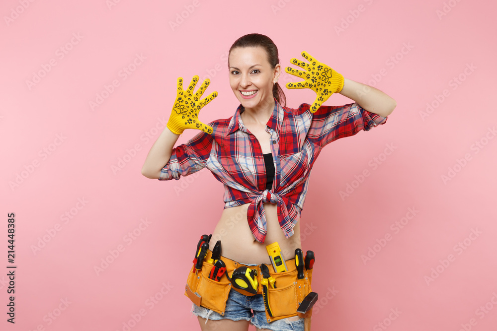 Strong young fun handyman woman in plaid shirt, denim shorts, yellow gloves, kit tools belt full of variety useful instruments isolated on pink background. Female doing male work. Renovation concept. Strong young fun handyman woman in plaid shirt, denim shorts, yellow gloves, kit tools belt full of variety useful instruments isolated on pink background. Female doing male work. Renovation concept.