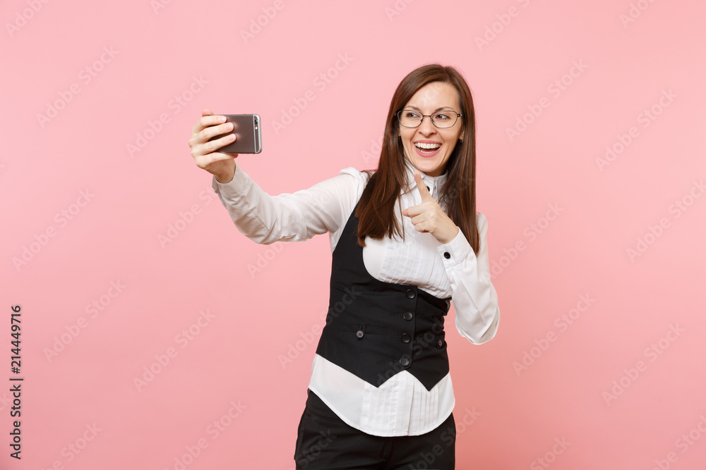 Young laughing business woman in glasses doing taking selfie shot on mobile phone pointing index finger isolated on pink background. Lady boss. Achievement career wealth. Copy space for advertisement. Young laughing business woman in glasses doing taking selfie shot on mobile phone pointing index finger isolated on pink background. Lady boss. Achievement career wealth. Copy space for advertisement.