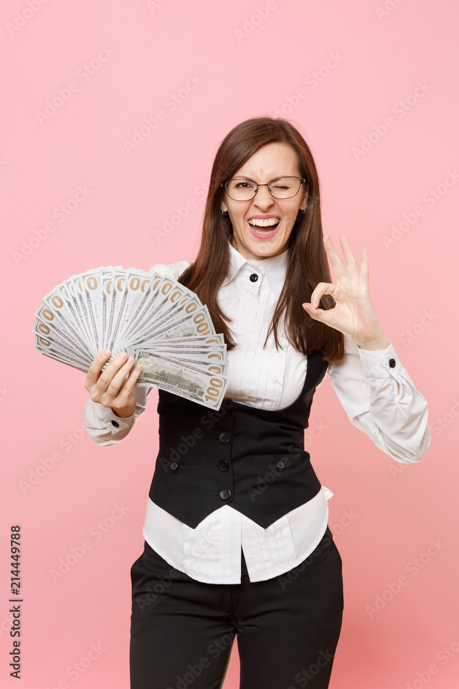 Young funny business woman in glasses blinking hold bundle lots of dollars, cash money showing OK sign isolated on pink background. Lady boss. Achievement career wealth. Copy space for advertisement. Young funny business woman in glasses blinking hold bundle lots of dollars, cash money showing OK sign isolated on pink background. Lady boss. Achievement career wealth. Copy space for advertisement.