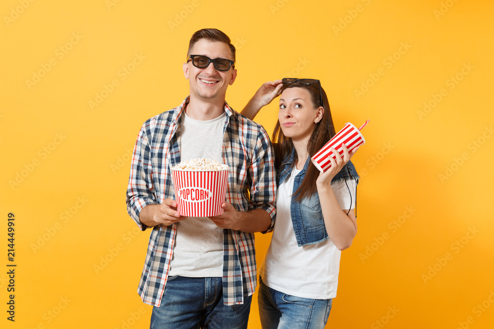Young smiling couple woman and man in 3d glasses watching movie film on date holding bucket of popcorn and plastic cup of soda or cola isolated on yellow background. Emotions in cinema concept. Young smiling couple woman and man in 3d glasses watching movie film on date holding bucket of popcorn and plastic cup of soda or cola isolated on yellow background. Emotions in cinema concept.