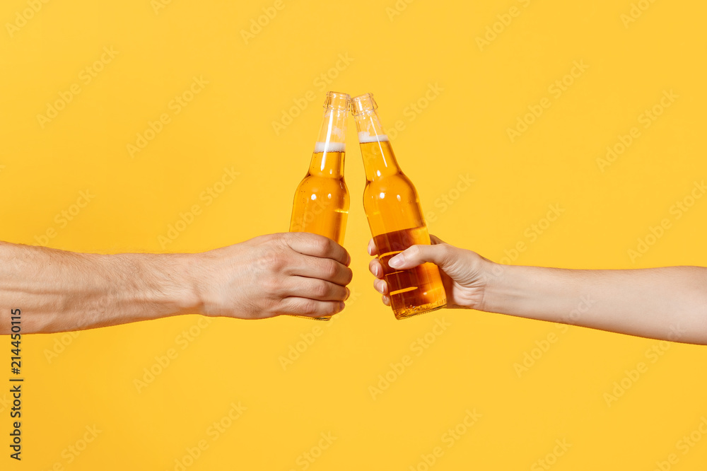 Close up cropped of woman and man two hands horizontal holding lager beer glass bottles and clinking isolated on yellow background. Sport fans cheer up. Friends leisure lifestyle concept. Copy space. - obrazy, fototapety, plakaty 
