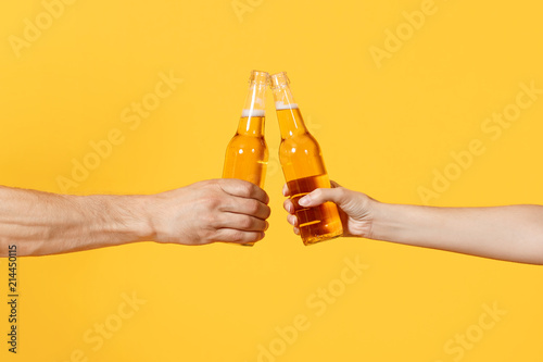 Fotobehang Close up cropped of woman and man two hands horizontal holding lager beer glass bottles and clinking isolated on yellow background