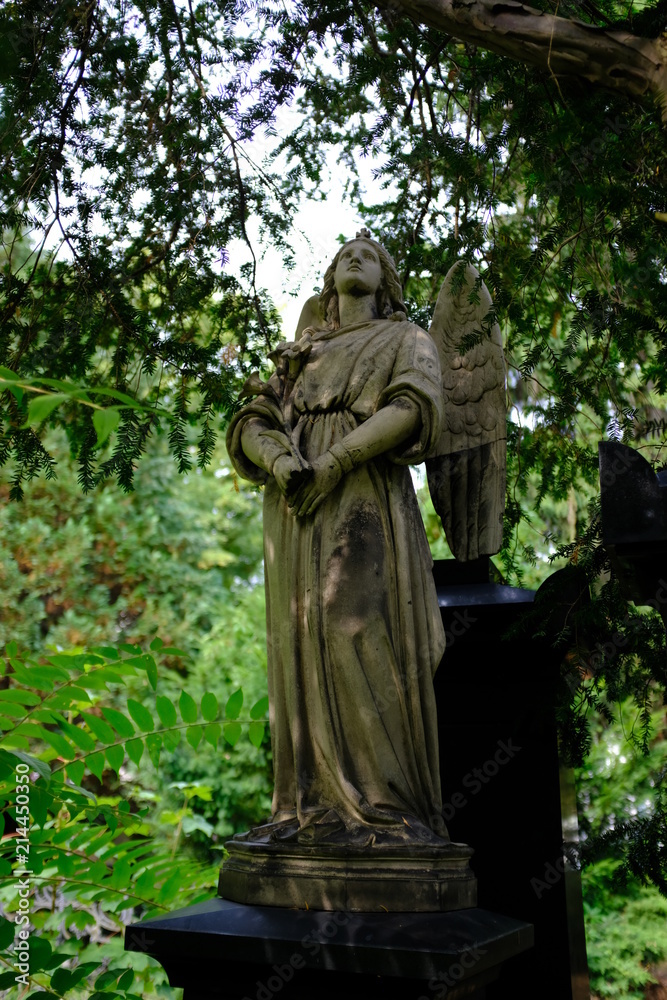 A weathered sandstone sculpture of an angel under trees. A cemetery in Berlin-Germany.
