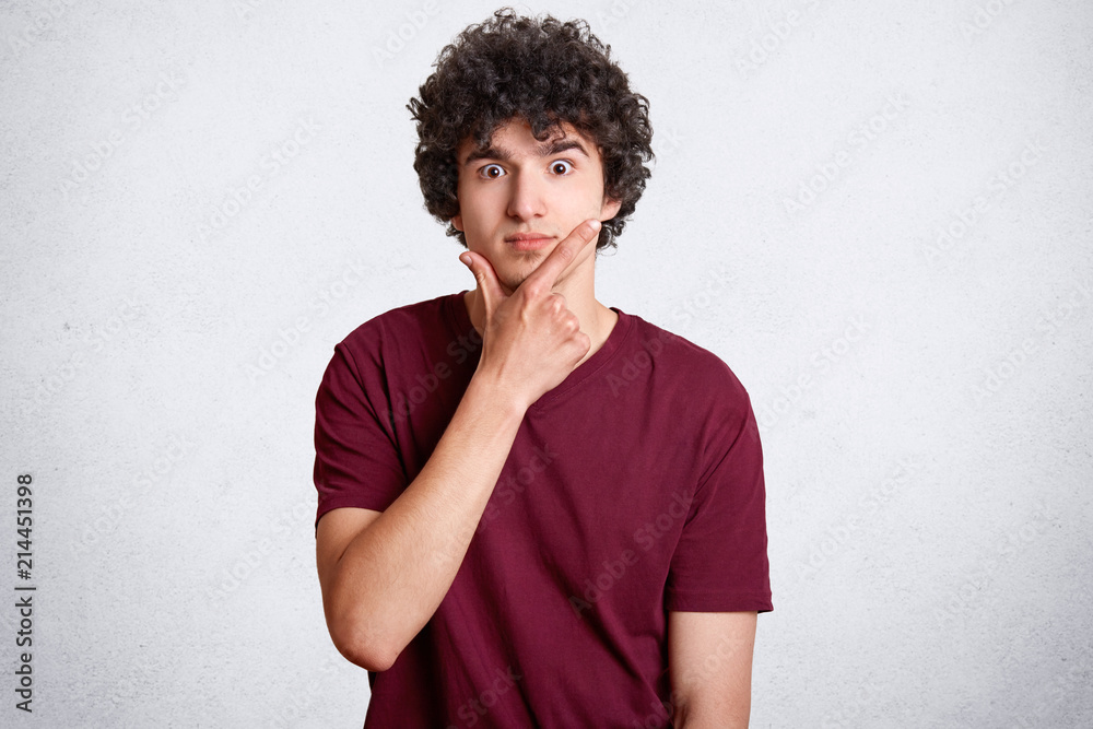 Surprised curly male holds chin and looks with shocked expression, has eyes full of disbelief, wears red casual t shirt, isolated over white concrete wall. People, emotions and reaction concept Surprised curly male holds chin and looks with shocked expression, has eyes full of disbelief, wears red casual t shirt, isolated over white concrete wall. People, emotions and reaction concept