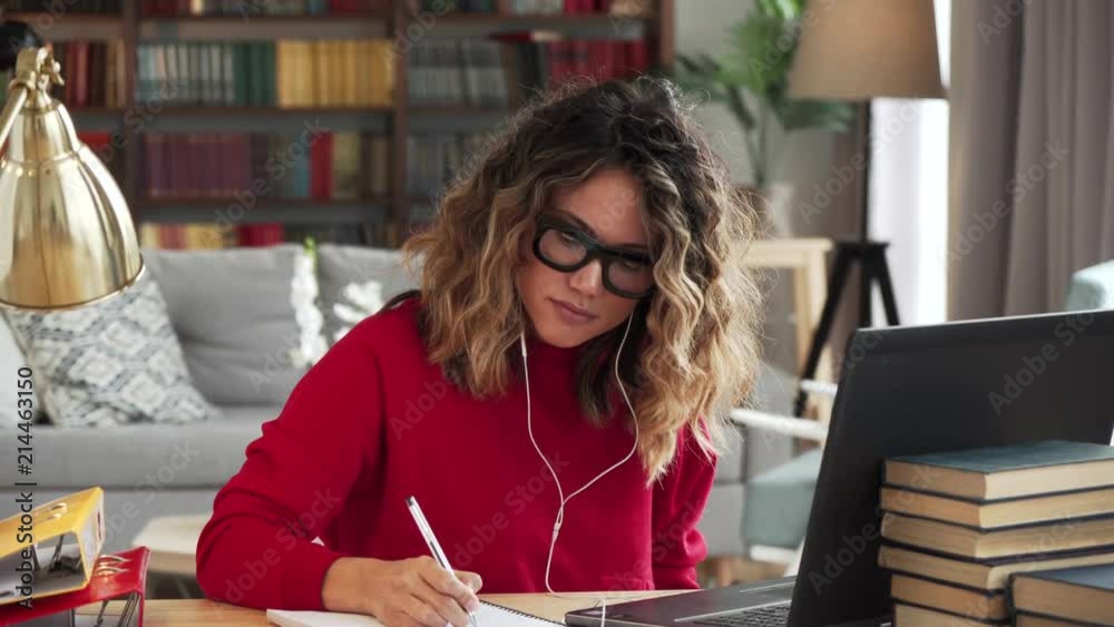 Beautiful curly woman in thirties of fair complexion in red jersey glasses and headphones studies online makes notes watches listens to course on laptop at the table with books in stylish room at home