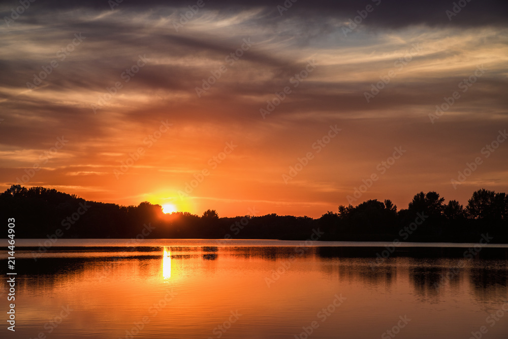 Setting sun over silhouettes of trees with reflection in lake