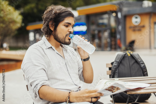 Attractive man sitting at street cafe, looking at the map and drinking water.
