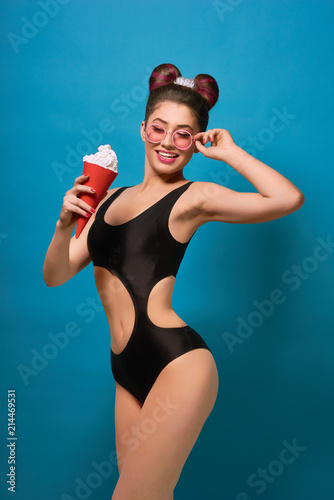 Smiling girl wearing black stylish swimwear posing with red whipped cream can. Having slim figure, fancy hairdress with a bow, day make up with pink lipstick. Standing on blue studio background.