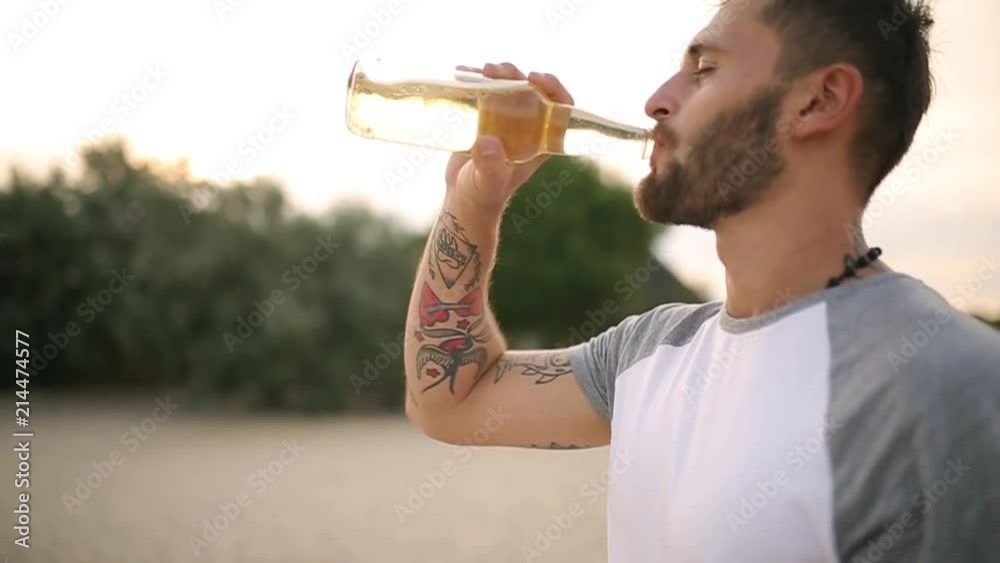 Young handsome tatooed bearded caucasian man drinking beer of glass bottle on the beach during sunset, steadycam shot, slow motion. Male quenches thirst with lemonade beverage at sandy sea shore.