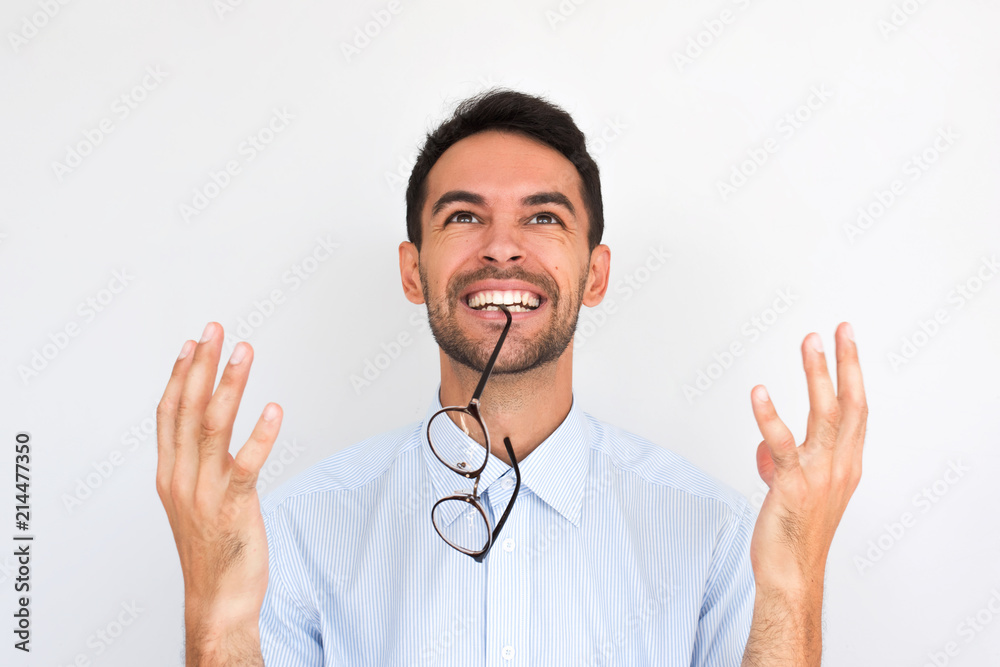 Closeup image of cheerful positive young Caucasian male with spectacles in mouth wins, gestures with happiness, wears blue shirt, posing against white studio wall. People, triumph and success concept Closeup image of cheerful positive young Caucasian male with spectacles in mouth wins, gestures with happiness, wears blue shirt, posing against white studio wall. People, triumph and success concept