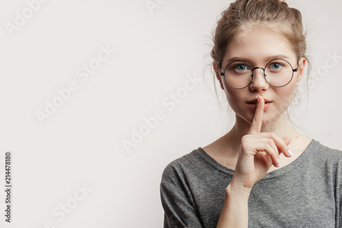 Hush. Close up portrait of young attractive female model in spectacles and misterious look asking for silence or secrecy with index finger on lips, shh hand gesture white background wall. photo