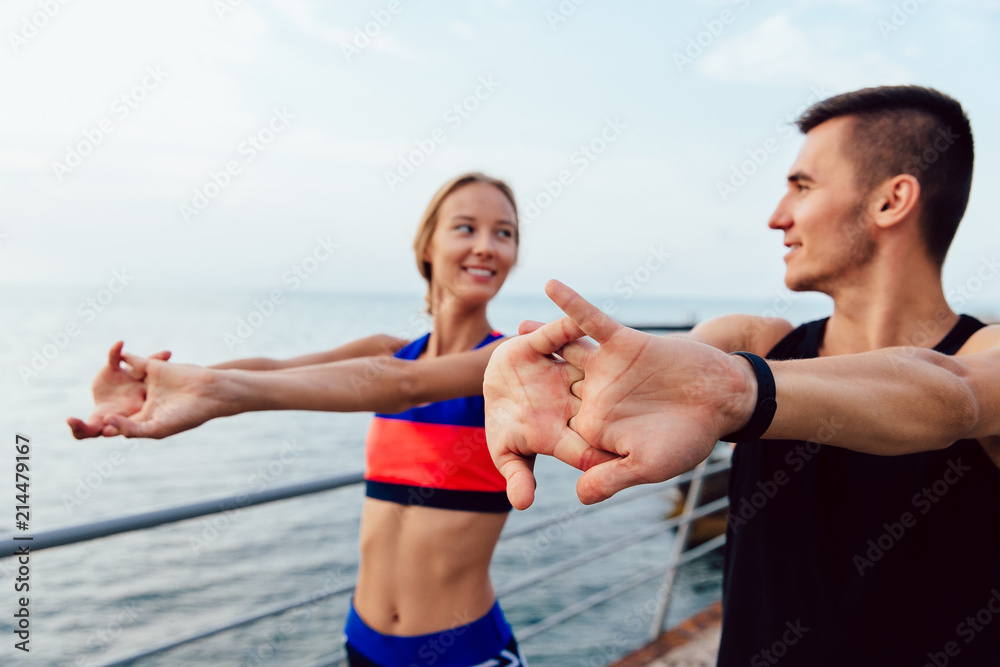 Sportive young couple doing stretching exercises for arms while working out on the quay, near the ocean. Healthy lifestyle. Sportive young couple doing stretching exercises for arms while working out on the quay, near the ocean. Healthy lifestyle.