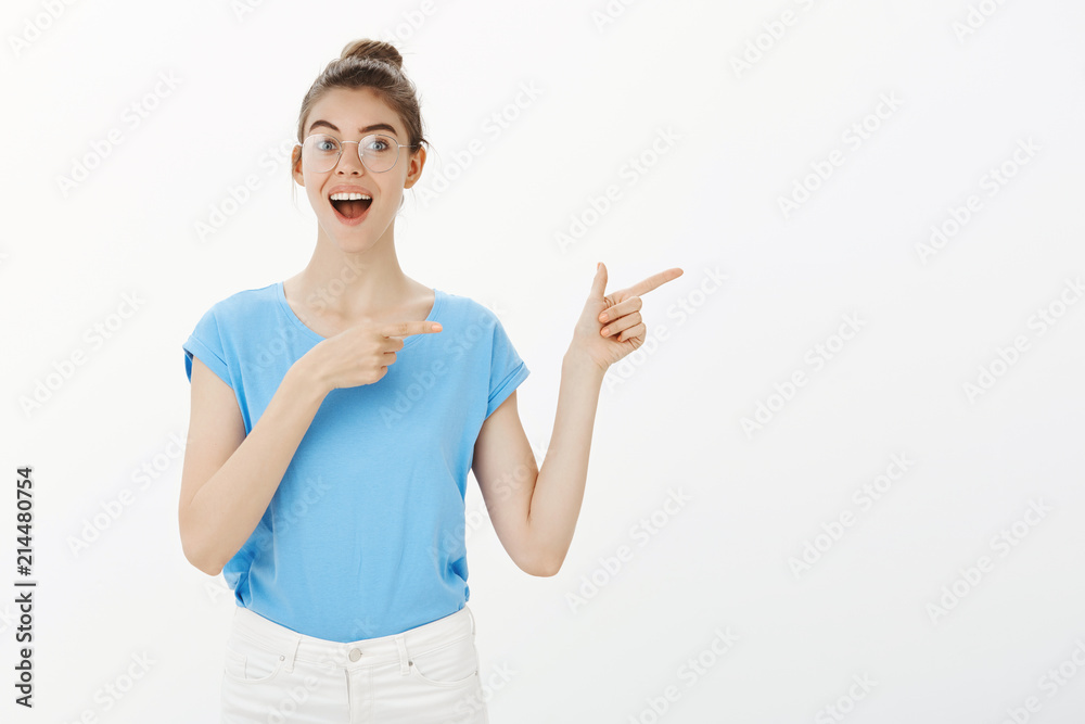 Portrait of good-looking happy young girl in glasses and bun, smiling broadly and pointing right with finger gun gestures, discussing great new place in town, suggesting friends to visit it Portrait of good-looking happy young girl in glasses and bun, smiling broadly and pointing right with finger gun gestures, discussing great new place in town, suggesting friends to visit it