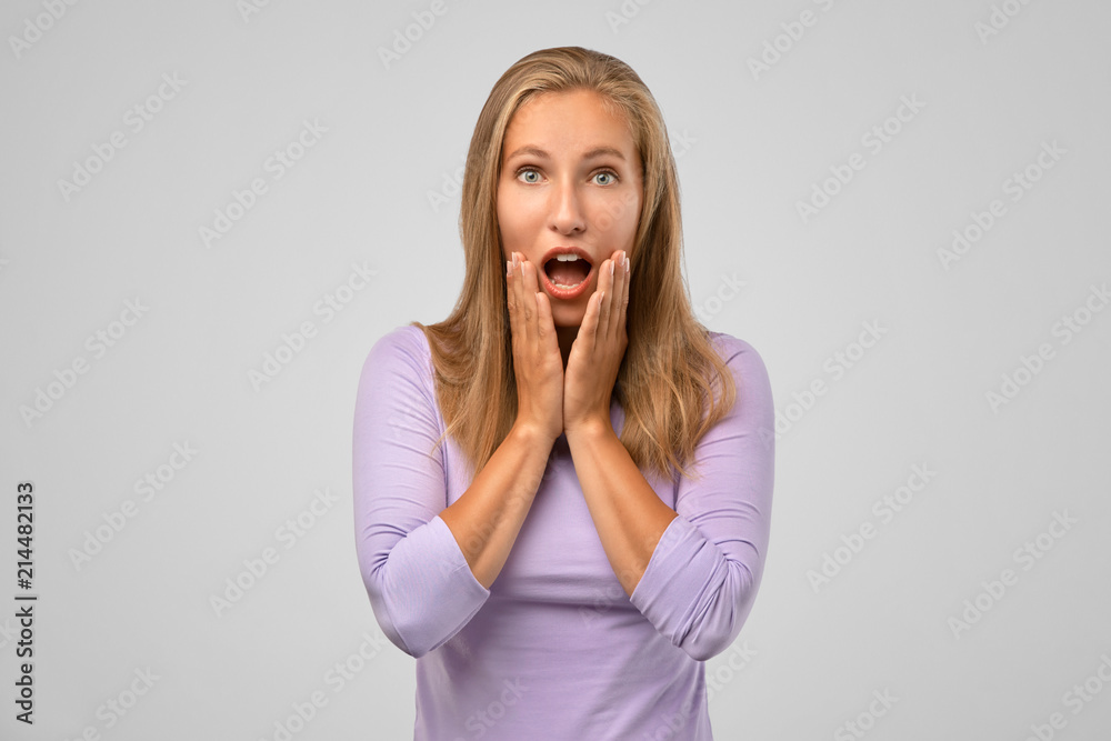Woman is shocked, looking with disbelief. Portrait of cute caucasian blonde standing partially crossing body with hand, holding palm on cheek and dropping jaw from stunning news, condemning friend Woman is shocked, looking with disbelief. Portrait of cute caucasian blonde standing partially crossing body with hand, holding palm on cheek and dropping jaw from stunning news, condemning friend