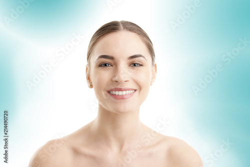 Portrait of a beautiful young woman with naked shoulders smiling while posing at isolated light blue background. Face beauty skin care. Natural make up