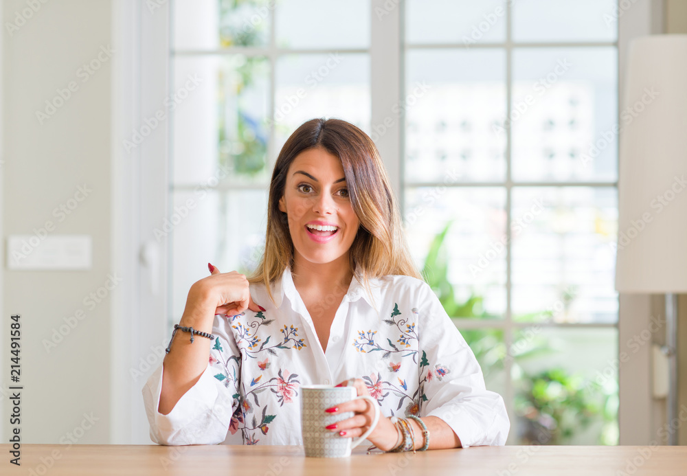 Young woman at home drinking coffee with surprise face pointing finger to himself Young woman at home drinking coffee with surprise face pointing finger to himself