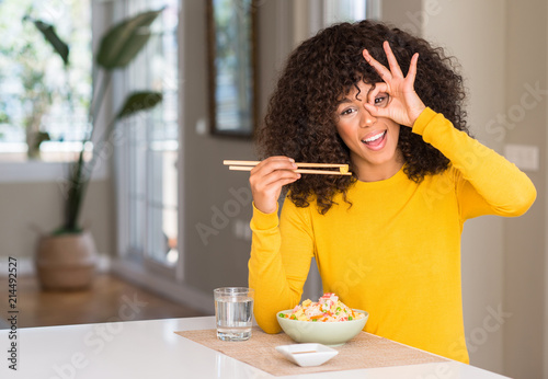 African american woman eating asian rice at home with happy face smiling doing ok sign with hand on eye looking through fingers