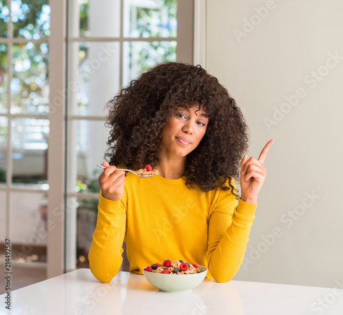 African american woman eating cereals, raspberries and blueberries very happy pointing with hand and finger to the side