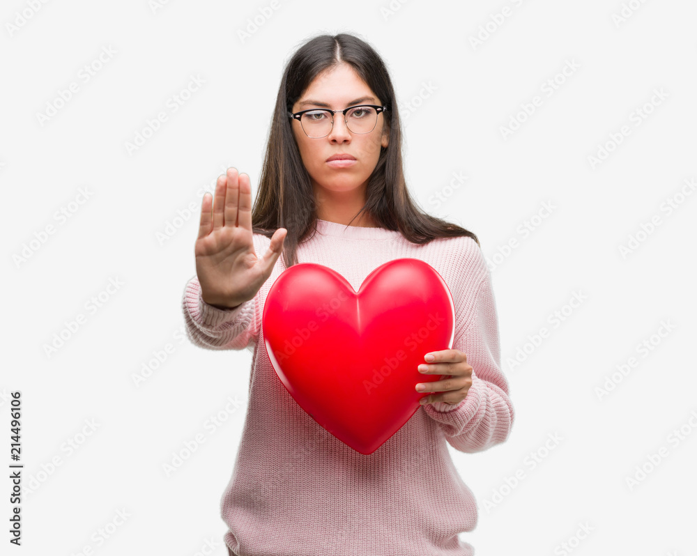 Young hispanic woman holding heart in love with open hand doing stop sign with serious and confident expression, defense gesture Young hispanic woman holding heart in love with open hand doing stop sign with serious and confident expression, defense gesture