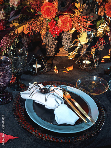 Table served in Halloween style. Bouquet with red flowers, decorative spiders, pumpkins with lettering and gothic vine glasses on black tablecloth. Holiday decorations.