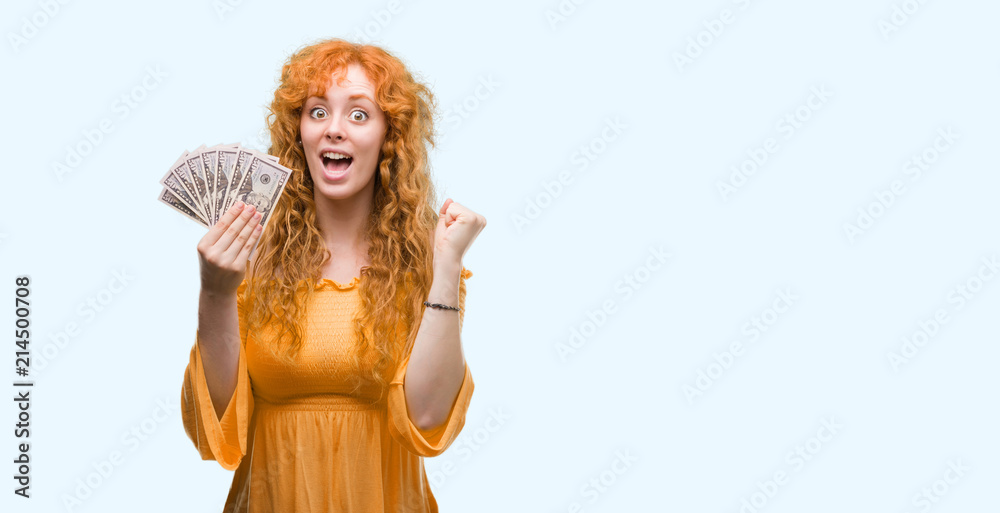 Young redhead woman holding dollars screaming proud and celebrating victory and success very excited, cheering emotion