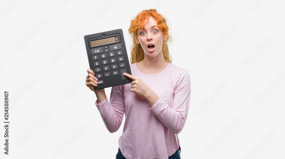 Young redhead woman holding big calculator scared in shock with a surprise face, afraid and excited with fear expression Young redhead woman holding big calculator scared in shock with a surprise face, afraid and excited with fear expression