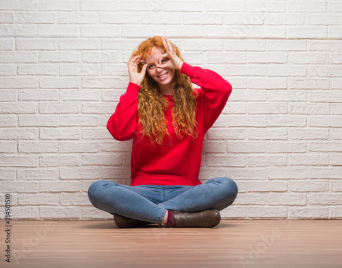 Young redhead woman sitting over brick wall doing ok gesture with hand smiling, eye looking through fingers with happy face. © Krakenimages.com
