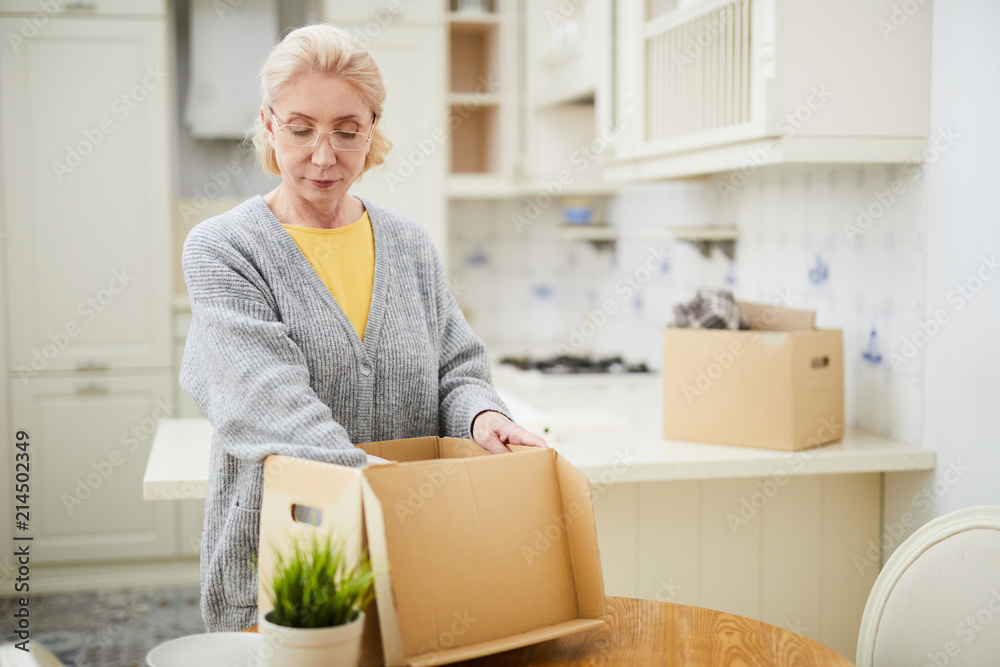 Blond aged woman putting things into carton box while standing by kitchen table Blond aged woman putting things into carton box while standing by kitchen table