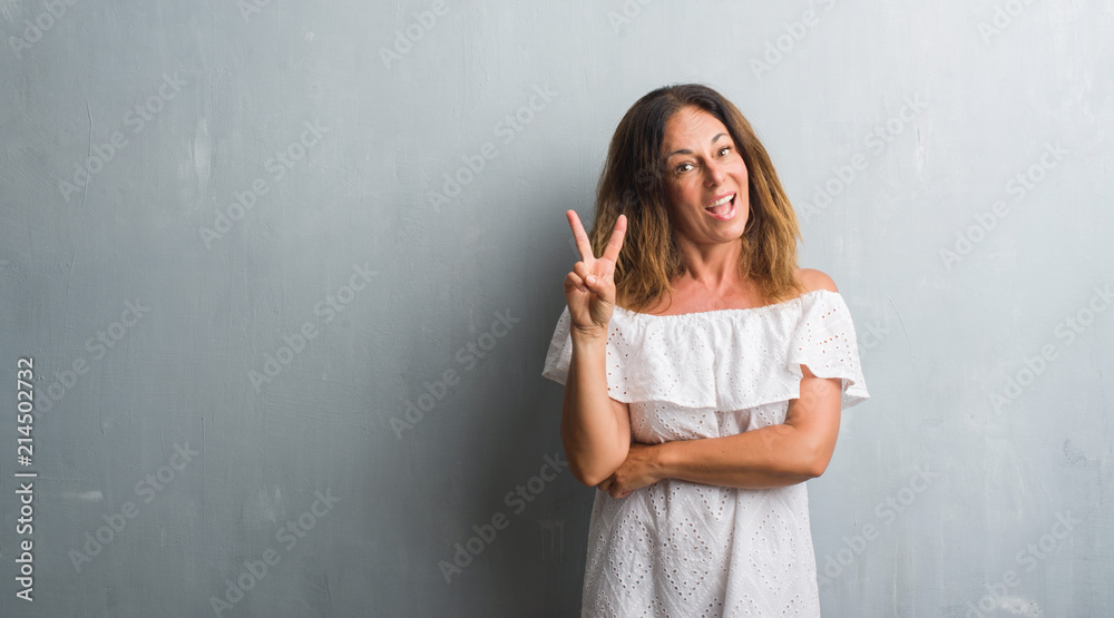 Middle age hispanic woman standing over grey grunge wall smiling with happy face winking at the camera doing victory sign. Number two. Middle age hispanic woman standing over grey grunge wall smiling with happy face winking at the camera doing victory sign. Number two.
