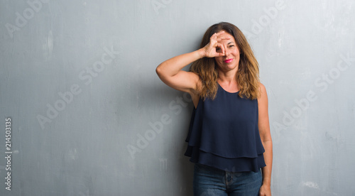 Middle age hispanic woman standing over grey grunge wall doing ok gesture with hand smiling, eye looking through fingers with happy face.