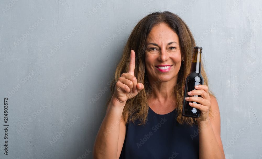Middle age hispanic woman standing over grey grunge wall holding beer bottle surprised with an idea or question pointing finger with happy face, number one Middle age hispanic woman standing over grey grunge wall holding beer bottle surprised with an idea or question pointing finger with happy face, number one
