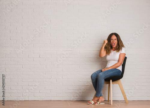 Middle age hispanic woman sitting on chair over white brick walll annoyed and frustrated shouting with anger, crazy and yelling with raised hand, anger concept