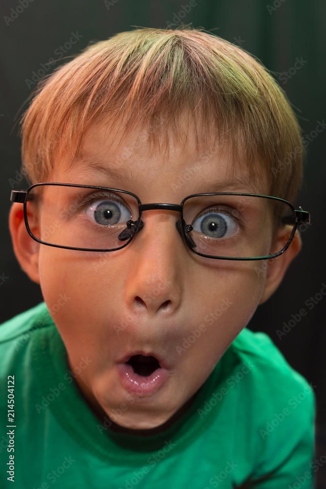 Casual boy with glasses with a stupid expression on his face, looks at the camera, against a dark background.