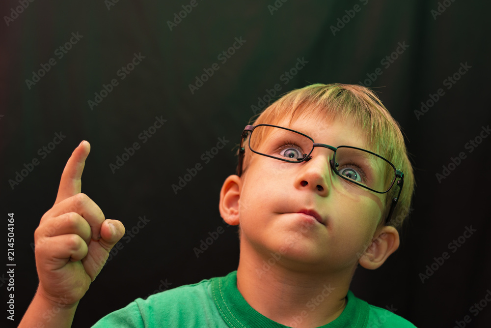 A casual boy with glasses raised his finger up and looks up, against a dark background.
