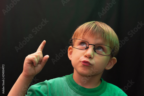 A casual boy with glasses raised his finger up and looks up  against a dark background.