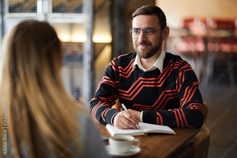 Obraz premium Young confident businessman in eyeglasses looking at colleague at meeting while making working notes