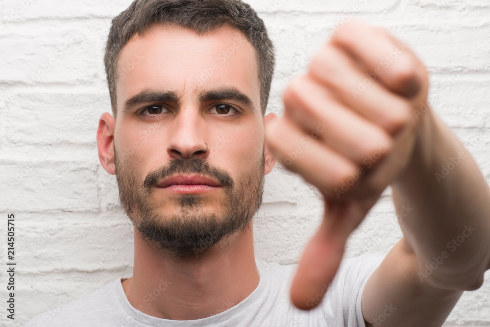 Obraz premium Young adult man standing over white brick wall with angry face, negative sign showing dislike with thumbs down, rejection concept