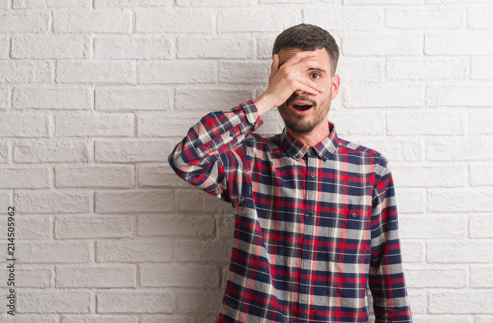 Young adult man standing over white brick wall peeking in shock covering face and eyes with hand, looking through fingers with embarrassed expression. Young adult man standing over white brick wall peeking in shock covering face and eyes with hand, looking through fingers with embarrassed expression.
