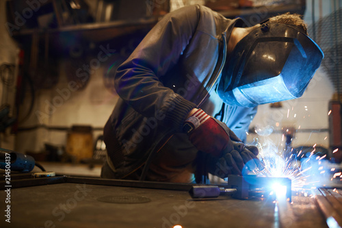 Young welder in protective mask leaning over workbench and welding iron workpieces