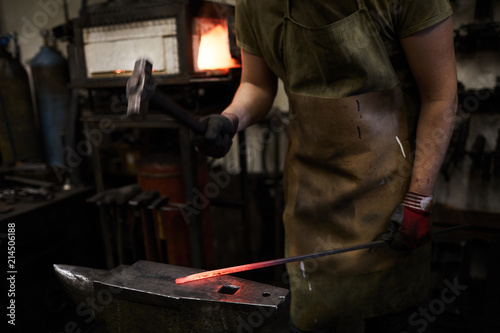 Blacksmith in uniform standing by iron anvil and forging hot metal workpiece with hammer