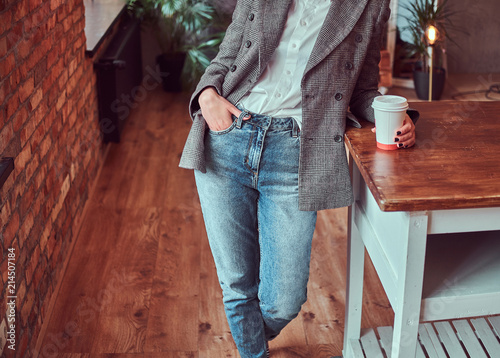 Cropped photo of a young woman dressed in a gray elegant jacket and jeans holding a cup of takeaway coffee while leaning on a table in a room with loft interior.